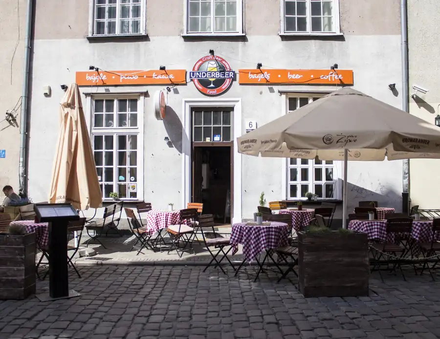 Biała kamienica Under Beer z pomarańczowym szyldem i stolikami przed wejściem